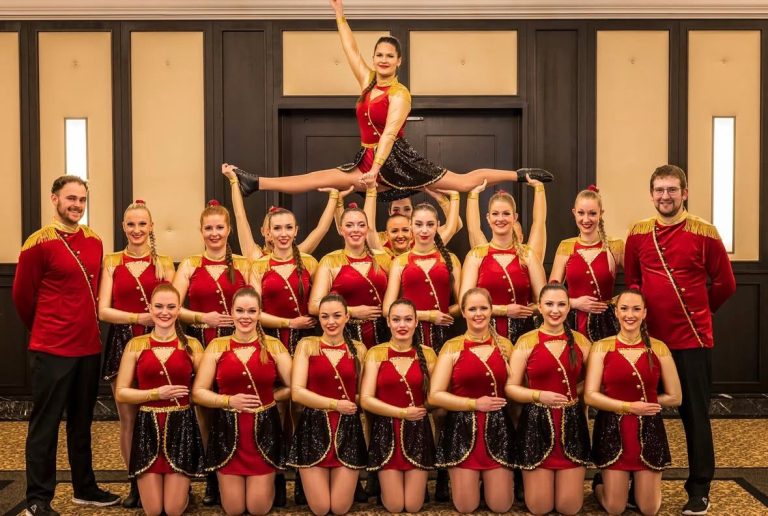 Gruppe von Tänzerinnen in rot-goldenen Kostümen, eine Tänzerin in der Luft schwebend.